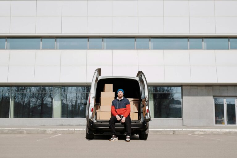 Man in a colorful jacket sitting by a van with boxes in an urban setting.